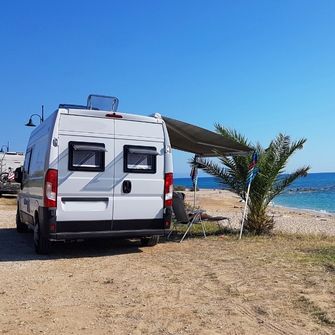 RV TOWING IN ORANGE COUNTY, CA. A SMALM MOTORHOME PARKED NEAR THE SAND  AT SAN CLEMENTE BEACH CAMP GROUNDS WITH A SHADE EXTENDING FROM THE RIGHT SIDE OF THE RV . A PALM TREE AND TWO CHAIRS ARE SITTING IN THE SAND UNDER THE SHADE IN FRONT OF THE OCEAN.
